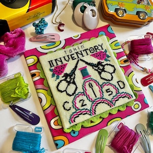 Peut inclure: Une collection d'articles d'artisanat et d'accessoires. Comprend un appareil photo jaune et noir, un collier doré avec un pendentif "C" rouge, une broderie avec le texte "takin' INVENTORY", et divers pinces à cheveux et bobines de fil.