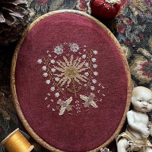 May include: A burgundy cross-stitch embroidery in an oval wooden hoop. The design features a central sunburst with gold and white details, surrounded by smaller floral elements. A spool of gold thread and a vintage doll are nearby.