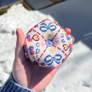 Puede incluir: Un cojín cuadrado hecho a mano con un fondo rosa claro y lazos azules bordados, acentos florales amarillos y rojos. Un pequeño botón azul claro está en el centro. El cojín se sostiene en una mano, con un fondo nevado.