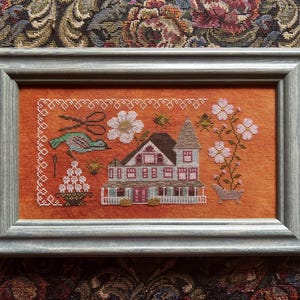 May include: A framed cross-stitch picture depicting a house with a tower, flowers, and birds, set against an orange backdrop. The artwork is enclosed in a grey wooden frame. The piece has a vintage, handcrafted feel, reminiscent of traditional needlework.