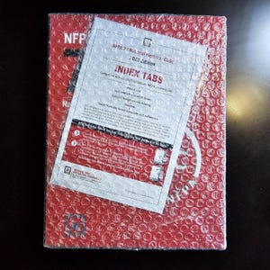 May include: A red book with the text "NFP" and "NFPA 70 National Electrical Code" is wrapped in bubble wrap. A white sheet with "INDEX TABS" and other text is on top of the book. The book is on a dark surface.