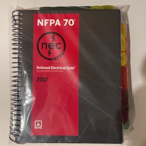 May include: A black spiral-bound book titled "NFPA 70 National Electrical Code 2017" with a red cover insert. The insert features the "nec" logo and the NFPA logo. The book is sealed in a clear plastic bag.