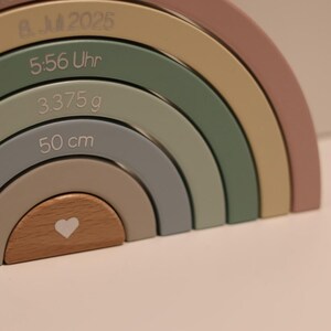 May include: A multi-colored wooden rainbow stacker toy featuring engraved text including 8. 2025, 5:56 Uhr, 3.375 g, and 50 cm. The arches are painted in muted tones of dusty rose, sage green, mustard yellow, and gray, with a natural wood center piece.
