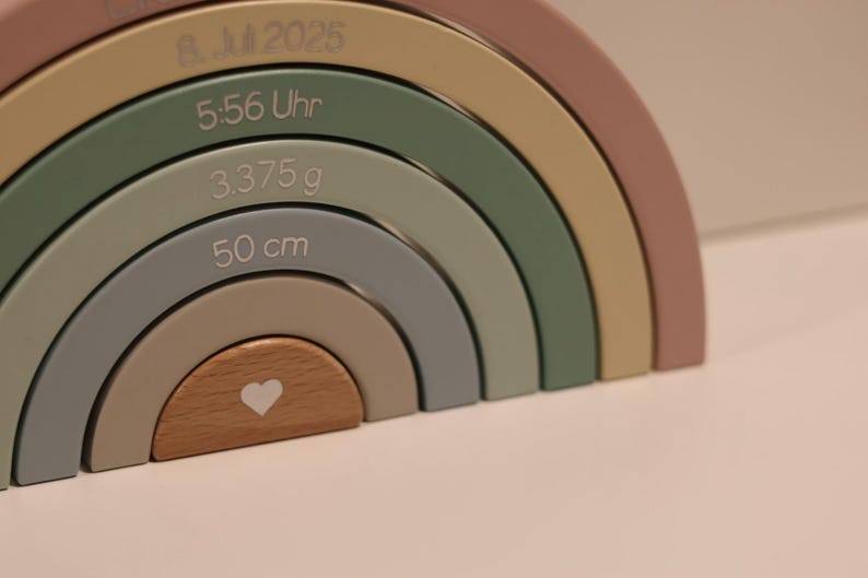 May include: A multi-colored wooden rainbow stacker toy featuring engraved text including 8. 2025, 5:56 Uhr, 3.375 g, and 50 cm. The arches are painted in muted tones of dusty rose, sage green, mustard yellow, and gray, with a natural wood center piece.