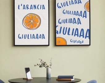 May include: Two framed art prints with black frames. The left print says "L'ARANCIA" above a drawing of an orange slice and "GIULIAAAA" below. The right print says "GIULIAAAA" multiple times with an orange slice illustration.