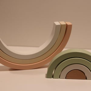 May include: Two sets of nested wooden arches. One set is arranged in a large upward-facing curve with muted peach, beige, and white tones. The second set is arranged in a smaller downward-facing arch with sage green, light gray, and brown wooden segments.