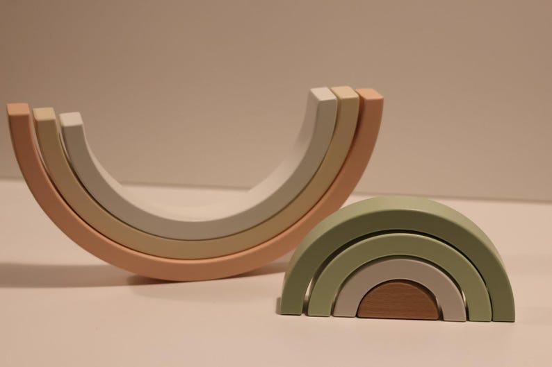 May include: Two sets of nested wooden arches. One set is arranged in a large upward-facing curve with muted peach, beige, and white tones. The second set is arranged in a smaller downward-facing arch with sage green, light gray, and brown wooden segments.