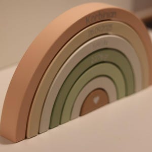May include: A wooden rainbow stacker toy featuring six nested arches in muted shades of peach, beige, and sage green. The arches are engraved with the name Korbinian, the date 26.09.2025, the time 04:19, the weight 2270gr, and a small heart icon.