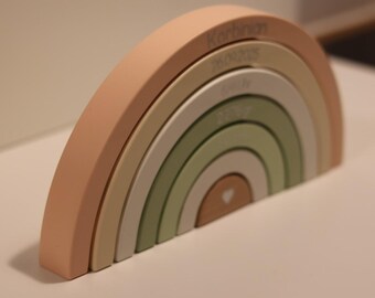 May include: A wooden rainbow stacker in muted pastel colors. The rainbow has layers of peach, beige, white, light green, and a brown base with a heart. The name "Korbinian" and the date "26.01.2020" are engraved on the rainbow.