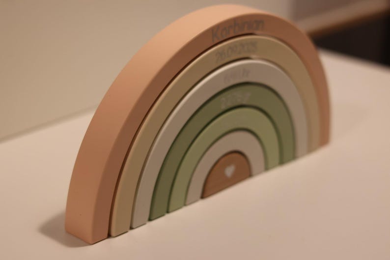 May include: A wooden rainbow stacker toy featuring six nested arches in muted shades of peach, beige, and sage green. The arches are engraved with the name Korbinian, the date 26.09.2025, the time 04:19, the weight 2270gr, and a small heart icon.