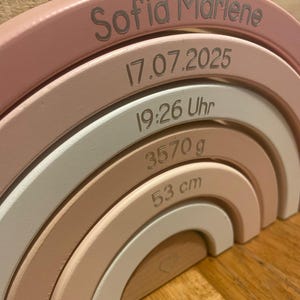 May include: A wooden rainbow stacker featuring five arched segments in shades of pink, white, and natural wood. The segments are engraved with the name Sofia Marlene, the date 17.07.2025, the time 19:26 Uhr, the weight 3570 g, and the length 53 cm.