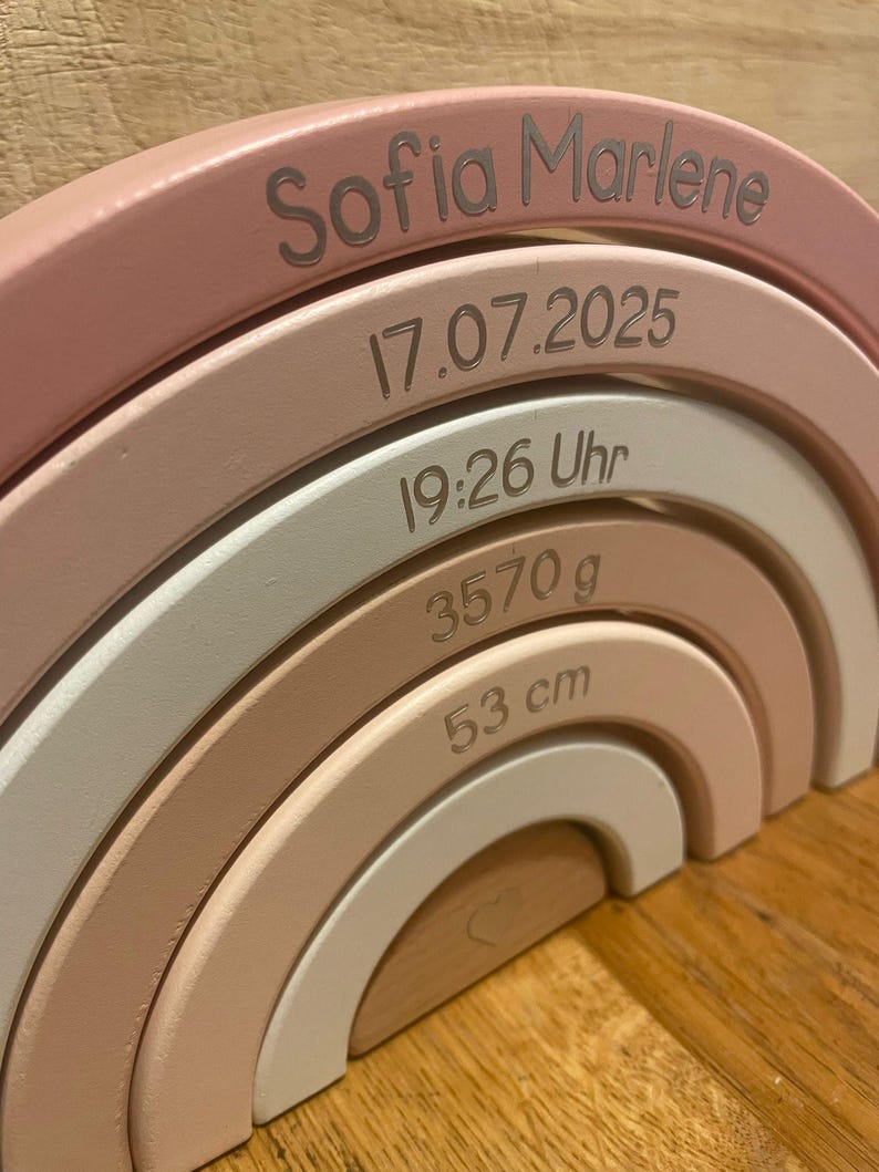 May include: A wooden rainbow stacker featuring five arched segments in shades of pink, white, and natural wood. The segments are engraved with the name Sofia Marlene, the date 17.07.2025, the time 19:26 Uhr, the weight 3570 g, and the length 53 cm.