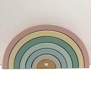 May include: A wooden rainbow stacking toy consisting of six nested arches in muted shades of dusty pink, pale yellow, sage green, blue-grey, and beige. The smallest central arch is natural wood with a small white heart printed on its front face.