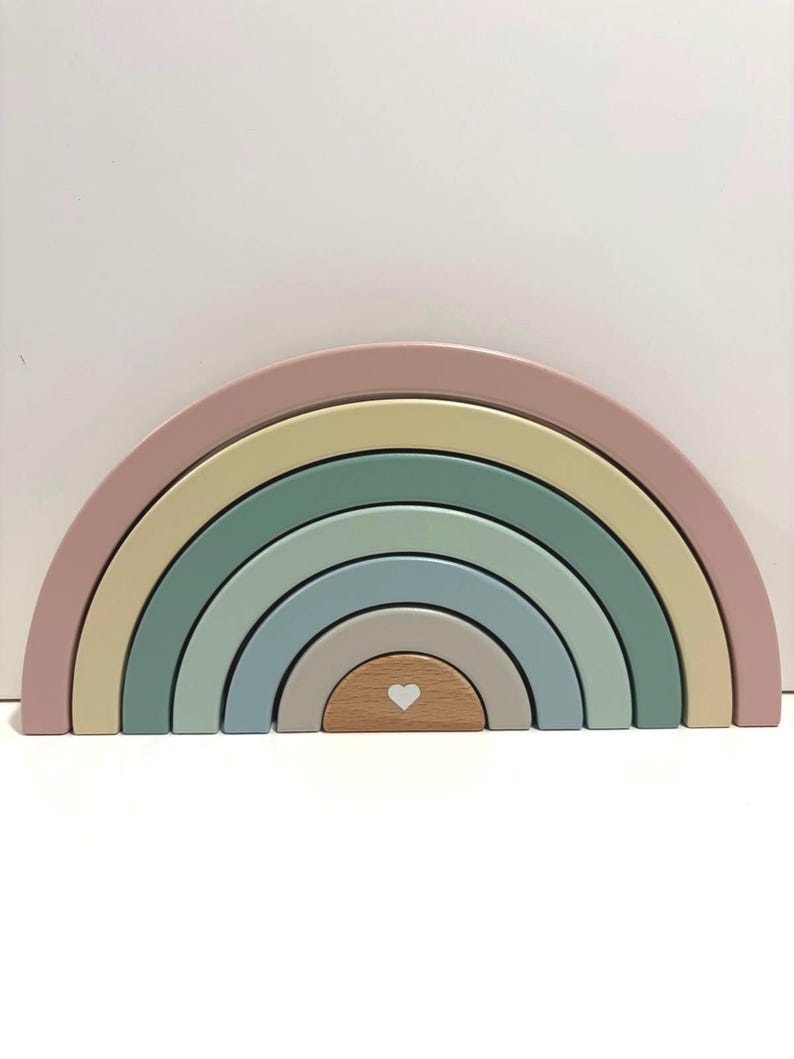 May include: A wooden rainbow stacking toy consisting of six nested arches in muted shades of dusty pink, pale yellow, sage green, blue-grey, and beige. The smallest central arch is natural wood with a small white heart printed on its front face.