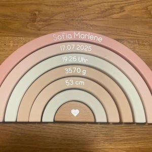 May include: A wooden rainbow stacker featuring six nested arches in shades of pink, beige, and sage green. The arches are printed with the name Sofia Marlene, the date 17.07.2025, the time 19:26 Uhr, the weight 3570 g, the length 53 cm, and a small heart.