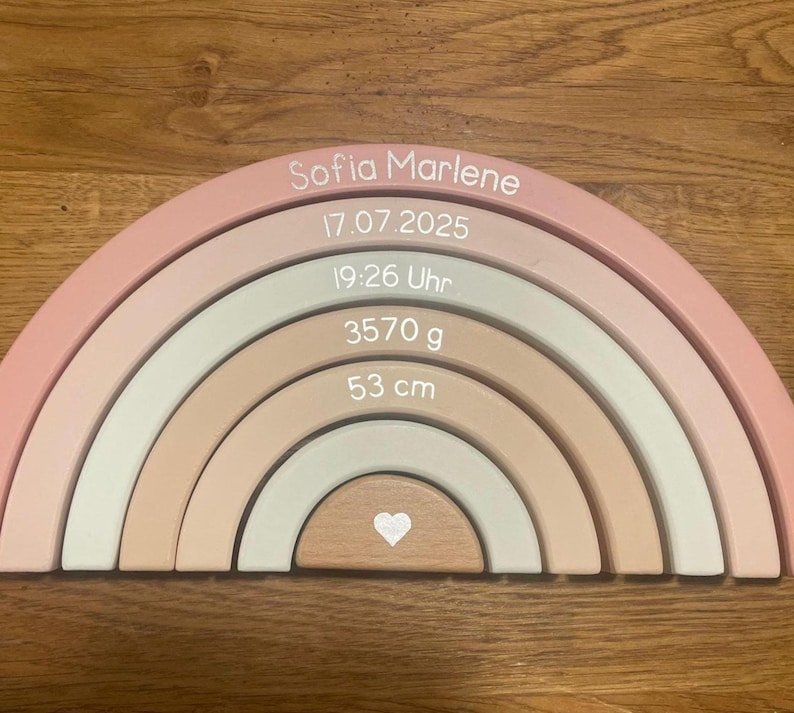 May include: A wooden rainbow stacker featuring six nested arches in shades of pink, beige, and sage green. The arches are printed with the name Sofia Marlene, the date 17.07.2025, the time 19:26 Uhr, the weight 3570 g, the length 53 cm, and a small heart.