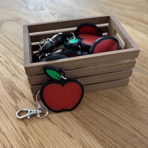 May include: A wooden crate filled with apple-shaped keychains. Each keychain features a red apple design with a black outline and a green leaf. The keychains have a silver clasp for easy attachment.
