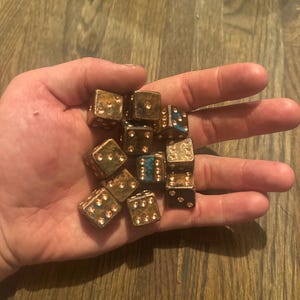 Op de afbeelding: Een verzameling dobbelstenen met een metallic gouden en zwarte afwerking. De dobbelstenen worden in een hand gehouden, waarbij hun verschillende zijden en puntarrangementen te zien zijn. Sommige dobbelstenen hebben blauwe accenten. De dobbelstenen worden waarschijnlijk gebruikt voor bordspellen.