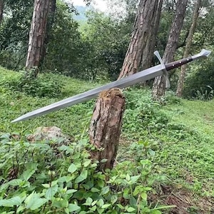 May include: A long, silver sword with a brown handle and crossguard rests on a tree stump. The sword is positioned diagonally, with the tip pointing towards the left. The background features lush green foliage and tall trees, creating a natural setting.