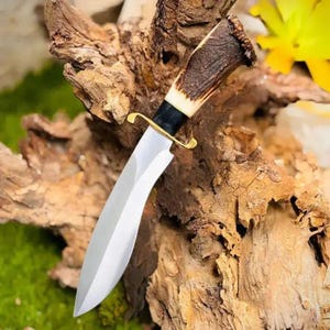 May include: A hunting knife with a curved, polished steel blade. The handle is made of brown antler with a black and gold guard. The knife is displayed against a backdrop of textured wood.