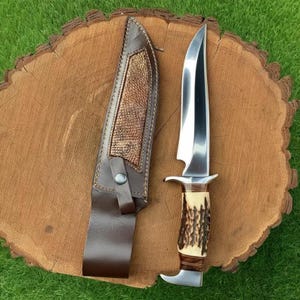 May include: A hunting knife with a polished steel blade and a handle made of bone and wood. The knife is next to a brown leather sheath with a decorative textured panel. The knife and sheath are on a wooden surface.