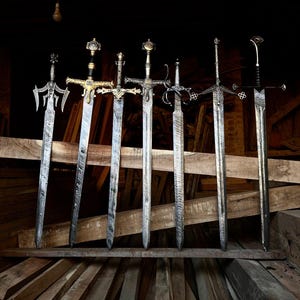 Könnte beinhalten: Sieben Metallschwerter mit kunstvollen Griffen und gemusterten Klingen in einer Reihe. Jedes Schwert hat ein einzigartiges Parierstangendesign, das von goldenen Akzenten bis zu komplexen silbernen Metallarbeiten reicht, alle mit einer Wellenstruktur.