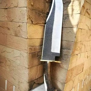 Puede incluir: Un cuchillo grande, hecho a mano, con una hoja pulida de color plateado y un mango oscuro y texturizado. El cuchillo tiene una virola de color latón y se exhibe contra una pared de piedra texturizada. La hoja mide unos 30 cm de largo.