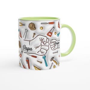 Puede incluir: Una taza de cer&aacute;mica blanca con asa e interior verde claro. La taza presenta un patr&oacute;n repetido de herramientas y la palabra "Papa" con un gr&aacute;fico de pu&ntilde;o. La taza es de tama&ntilde;o est&aacute;ndar.