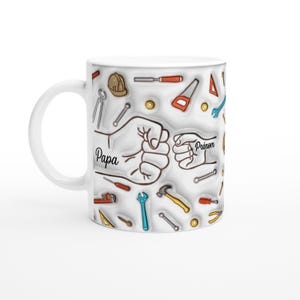 Puede incluir: Taza de cer&aacute;mica blanca con un dise&ntilde;o de herramientas y dos pu&ntilde;os chocando. La palabra "Papa" est&aacute; escrita en la taza. La taza tiene un asa blanca y un interior blanco. Las herramientas son de varios colores.