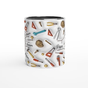 Puede incluir: Una taza de cer&aacute;mica blanca con interior negro, cubierta con un patr&oacute;n repetido de herramientas manuales en varios colores. La taza tiene la palabra "Papa" y la palabra "Pr&eacute;nom" impresa.