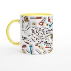 Puede incluir: Una taza de cer&aacute;mica blanca con asa e interior amarillos. La taza presenta un dise&ntilde;o de herramientas y pu&ntilde;os, con la palabra "Papa" y "Pr&eacute;nom" en texto negro. Las herramientas son de varios colores.
