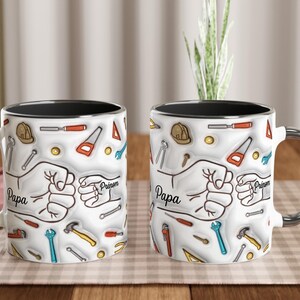 Puede incluir: Dos tazas de cer&aacute;mica blanca con bordes negros y asas plateadas. Cada taza presenta un dise&ntilde;o de herramientas, un pu&ntilde;o y la palabra "Papa". Las tazas est&aacute;n decoradas con ilustraciones de martillos, sierras y llaves.