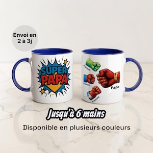 Könnte beinhalten: Zwei weiße Keramikbecher mit blauen Henkeln und Rändern. Ein Becher zeigt den Text "SUPER PAPA" im Comic-Stil, der andere Illustrationen von Superhelden-Fäusten. Die Becher haben den Text "Papa" und "Prenom".