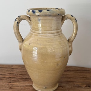 May include: A beige ceramic vase with two side handles and a wide rim. The vase features a glossy glaze finish with visible blue paint splatters along the rim and handles, showing a rounded body that tapers toward a narrow base.