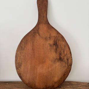 May include: A rustic, wooden cutting board with a handle. The board is oval-shaped with a long, slender handle. The wood has a warm, brown tone with visible grain and some darker areas, suggesting age and use. It rests on a wooden surface.
