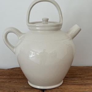 May include: A glossy off-white ceramic teapot featuring a rounded body, a side handle, a short spout, and a high arched handle extending over the top. The lid is centered with a small knob handle, and the entire piece has a smooth, reflective finish.