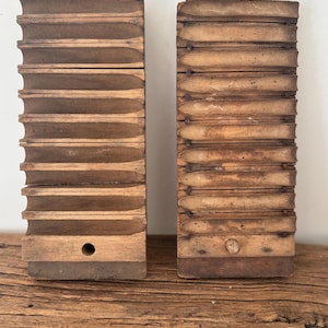 May include: Two rectangular wooden cigar molds standing upright, each featuring ten horizontal, semi-cylindrical grooves for shaping cigars. The left mold has a circular hole at the base, while the right mold shows a wooden plug in the same position.