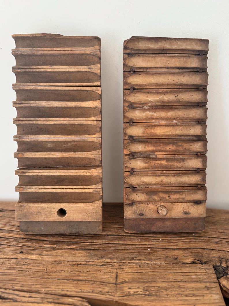 May include: Two rectangular wooden cigar molds standing upright, each featuring ten horizontal, semi-cylindrical grooves for shaping cigars. The left mold has a circular hole at the base, while the right mold shows a wooden plug in the same position.