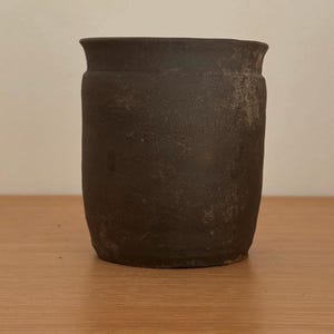 Puede incluir: Una maceta de cerámica marrón oscuro, cilíndrica, con un borde ligeramente ensanchado. La maceta tiene una superficie texturizada y parece hecha a mano. La maceta está sobre una superficie de madera.
