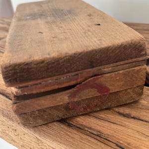 May include: A rectangular wooden box with a weathered, light brown finish and visible wood grain. The lid features a small, faded red circular stamp on the side edge. The construction appears simple with visible seams between the lid and the base.