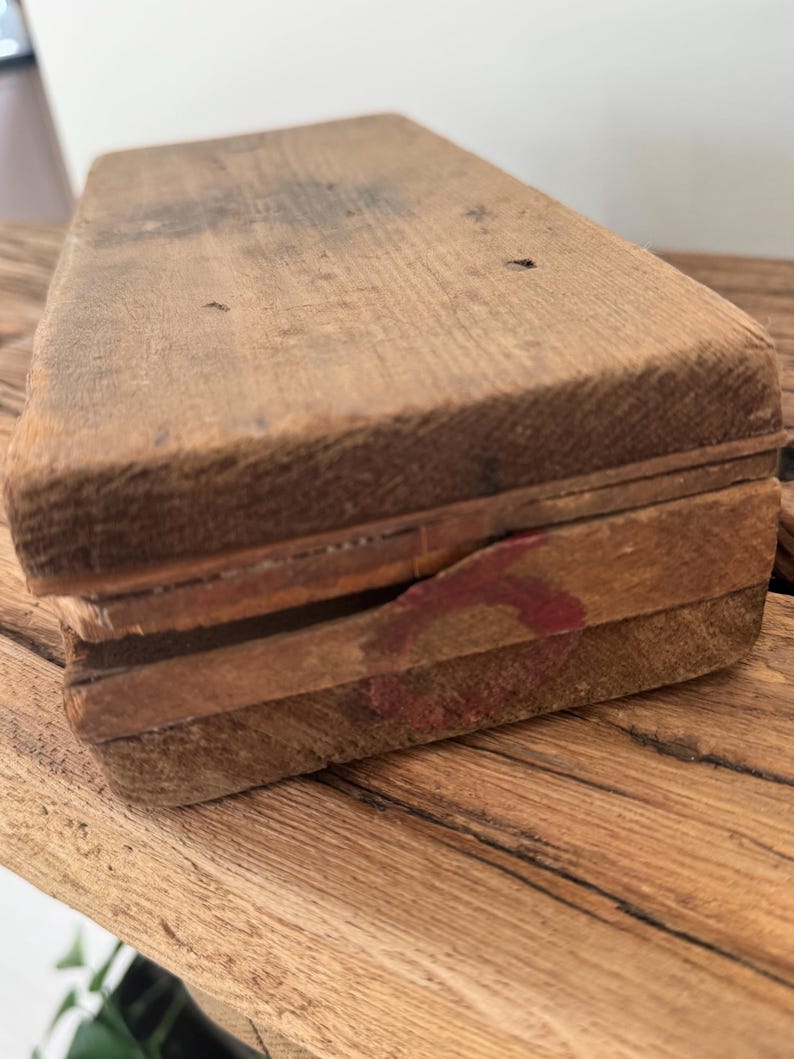 May include: A rectangular wooden box with a weathered, light brown finish and visible wood grain. The lid features a small, faded red circular stamp on the side edge. The construction appears simple with visible seams between the lid and the base.