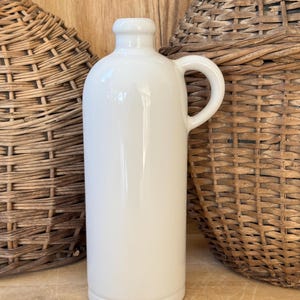 May include: A white ceramic bottle with a handle, set against a backdrop of woven baskets and a wooden surface. The bottle has a smooth, glossy finish and a rounded base. The handle is curved, and the opening is small.