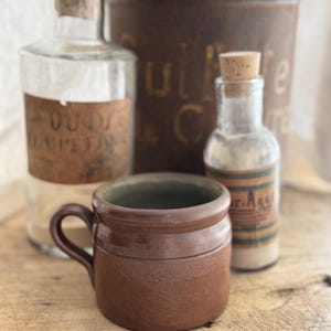 May include: A brown ceramic mug with a handle sits on a wooden surface. Behind it are glass bottles with cork stoppers and labels, and a tall, rusty metal container. The scene evokes a vintage, rustic aesthetic.