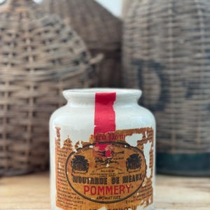 Puede incluir: Un tarro de cerámica blanco vintage de mostaza Moutarde de Meaux Pommery con una franja roja. La etiqueta está envejecida y parcialmente rasgada, con texto en francés. El tarro tiene una capacidad de 500 ml. Otras botellas tejidas están en el fondo.
