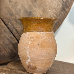 May include: A small, antique-style pottery vase with a rounded body and a flared, brown rim. The vase has a light brown base and a darker brown rim. The surface shows signs of age and wear, with visible texture and imperfections, giving it a rustic appearance.