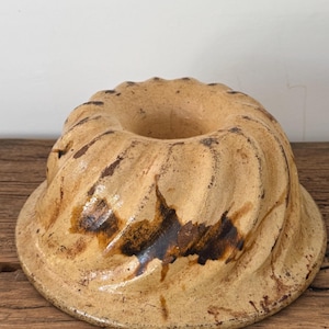 May include: A vintage, tan-colored Bundt cake pan with a fluted design. The ceramic pan has a circular shape with a central hole and a wide, slightly raised base. Dark brown markings add visual interest to the exterior.