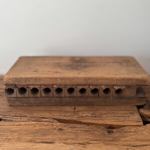 May include: A rectangular wooden cigar mold with a natural brown finish, featuring ten circular channels for shaping cigars. The side of the mold is stamped with the text 'MOLD' and 'USA' in a clear, dark font, showing visible wood grain texture.