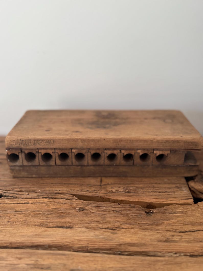May include: A rectangular wooden cigar mold with a natural brown finish, featuring ten circular channels for shaping cigars. The side of the mold is stamped with the text 'MOLD' and 'USA' in a clear, dark font, showing visible wood grain texture.