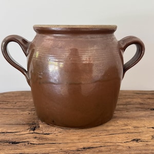 May include: A brown ceramic crock with two curved handles. The antique pot has a wide opening and a slightly lighter rim. The stoneware vessel is displayed on a wooden surface.