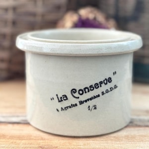 May include: A cream-colored ceramic jar with a rounded rim and a cylindrical body. The jar has the text "La Conserve" in black script, along with other text and the number "1/2". The jar is likely for food storage.
